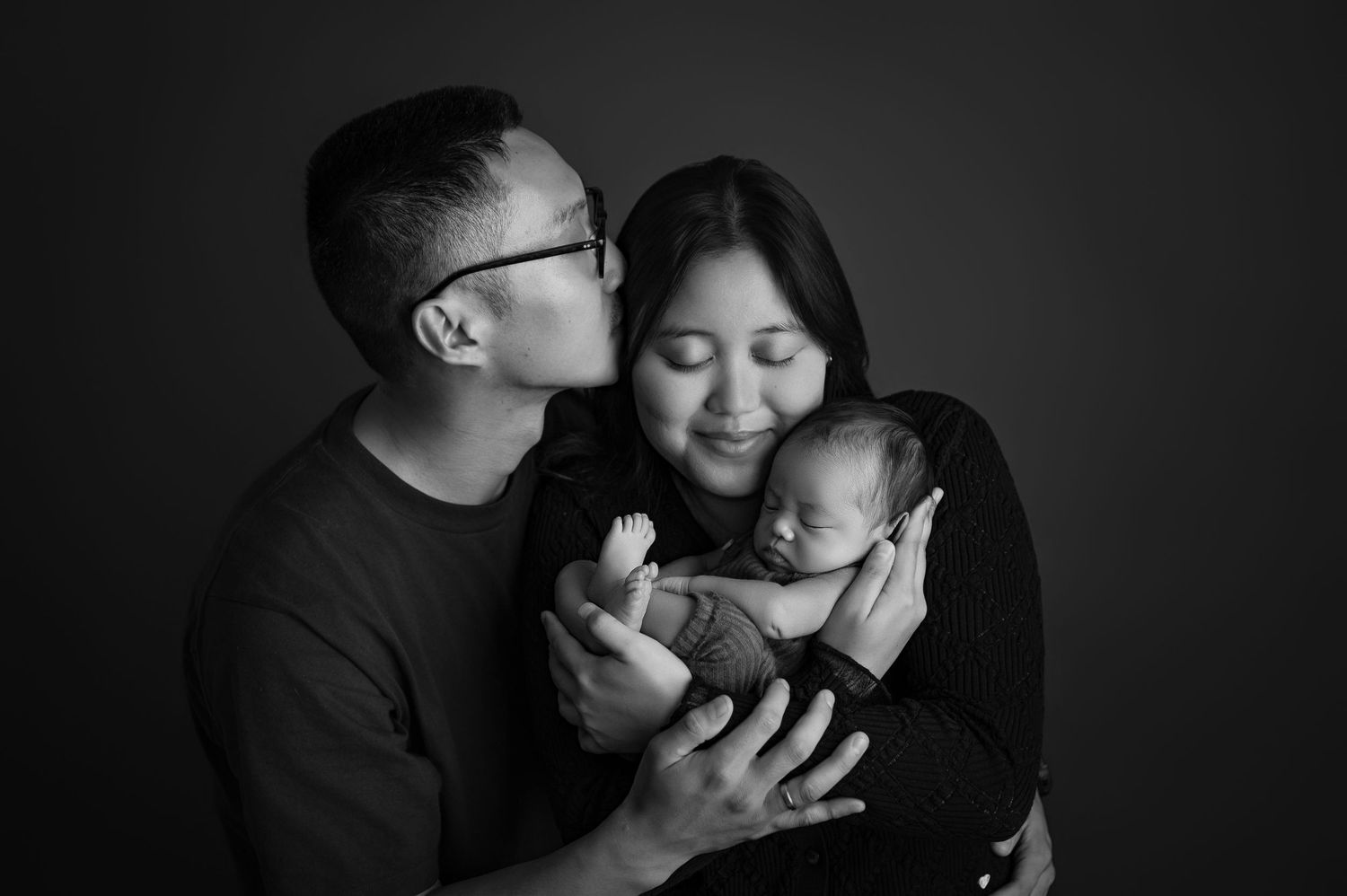 Black and white photo of parents lovingly holding their newborn baby, father kissing mother's cheek.