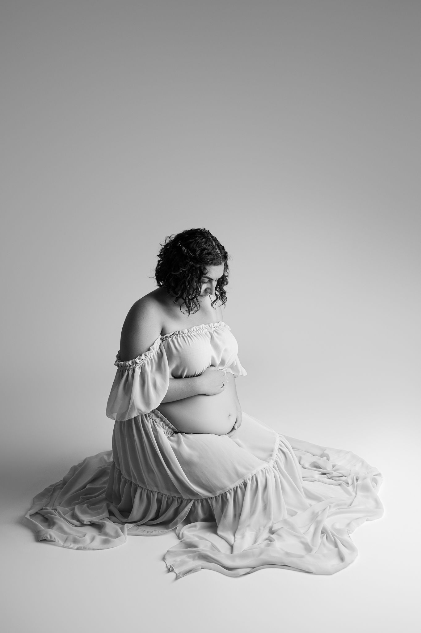 Black and white maternity photo of a pregnant woman in an off-shoulder gown sitting gracefully on a studio floor.