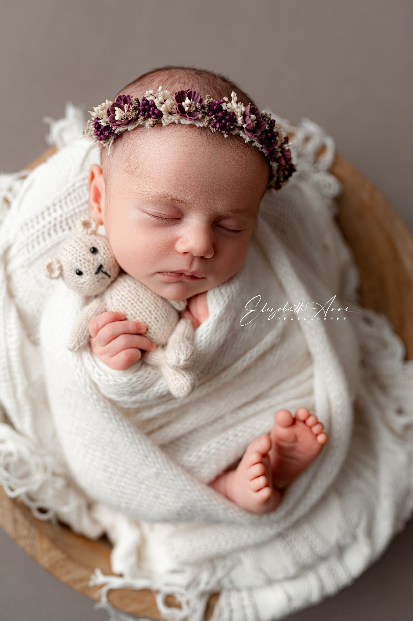 Sleeping newborn wrapped in cozy white knitted blanket wearing floral crown and cuddling small teddy bear.
