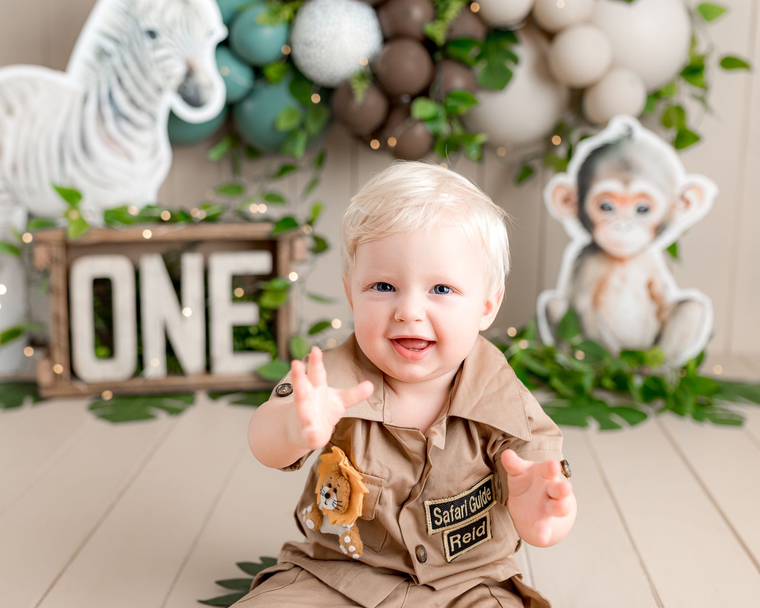 A small child in a tan outfit sits in front of a decorated backdrop with greenery and white accents.