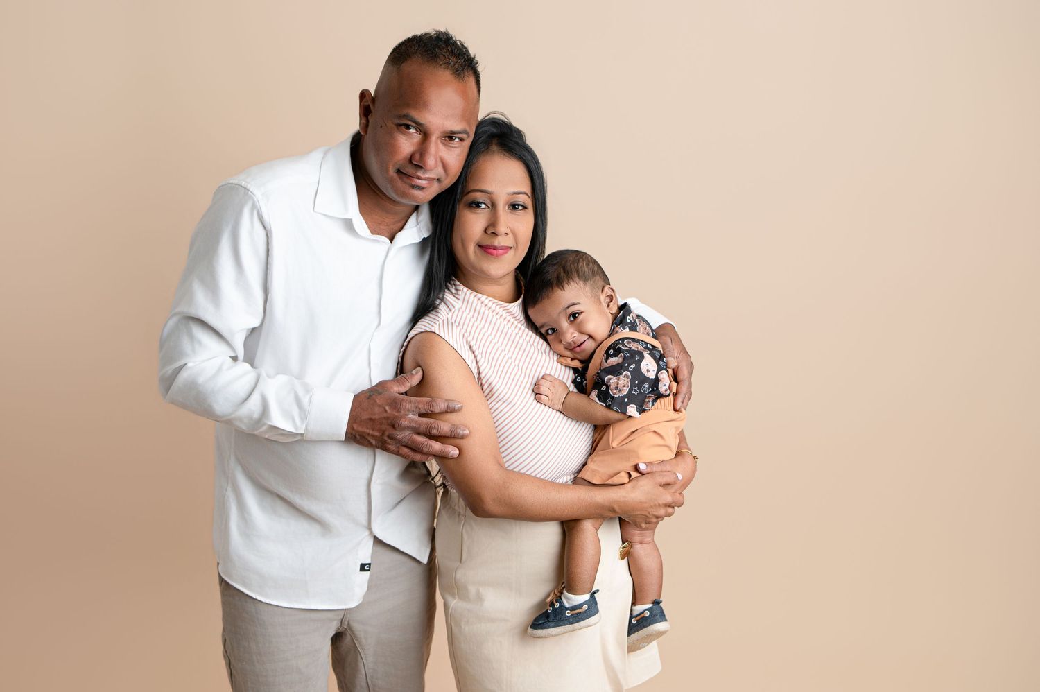 A smiling family of three poses together, with parents holding their baby boy, against a warm beige studio background.