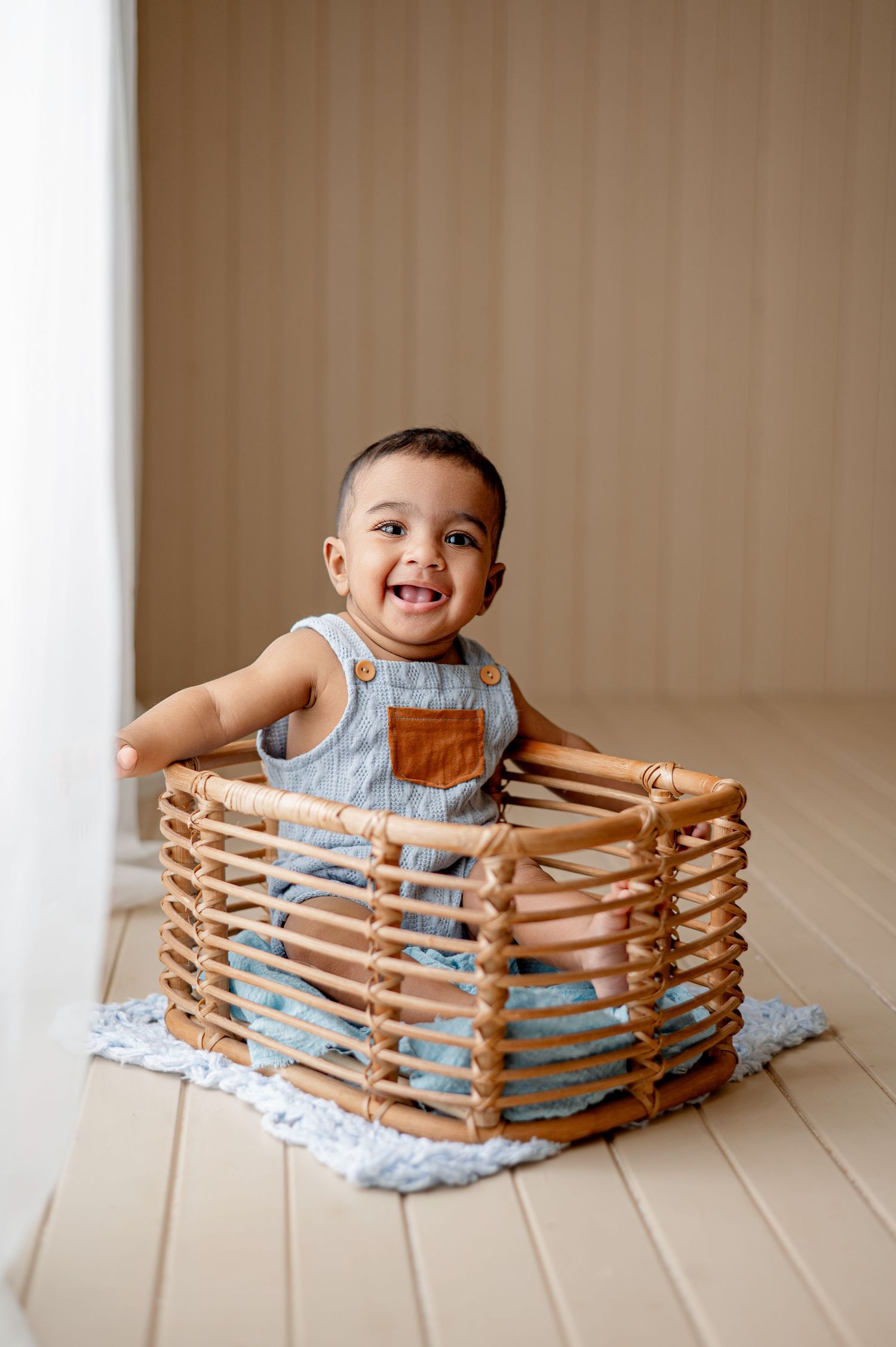 Smiling baby boy in denim overalls sitting inside a wicker basket on a light blue blanket in a bright room.