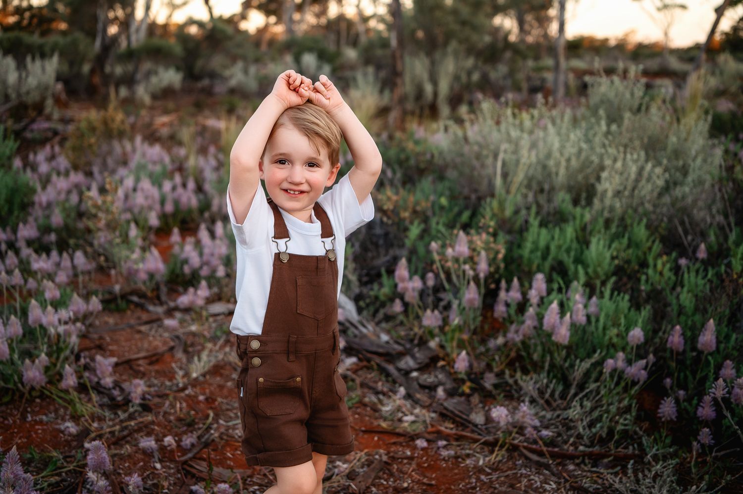 Young boy in brown overalls poses happily among pink wildflowers in an Australian outback landscape at golden hour.