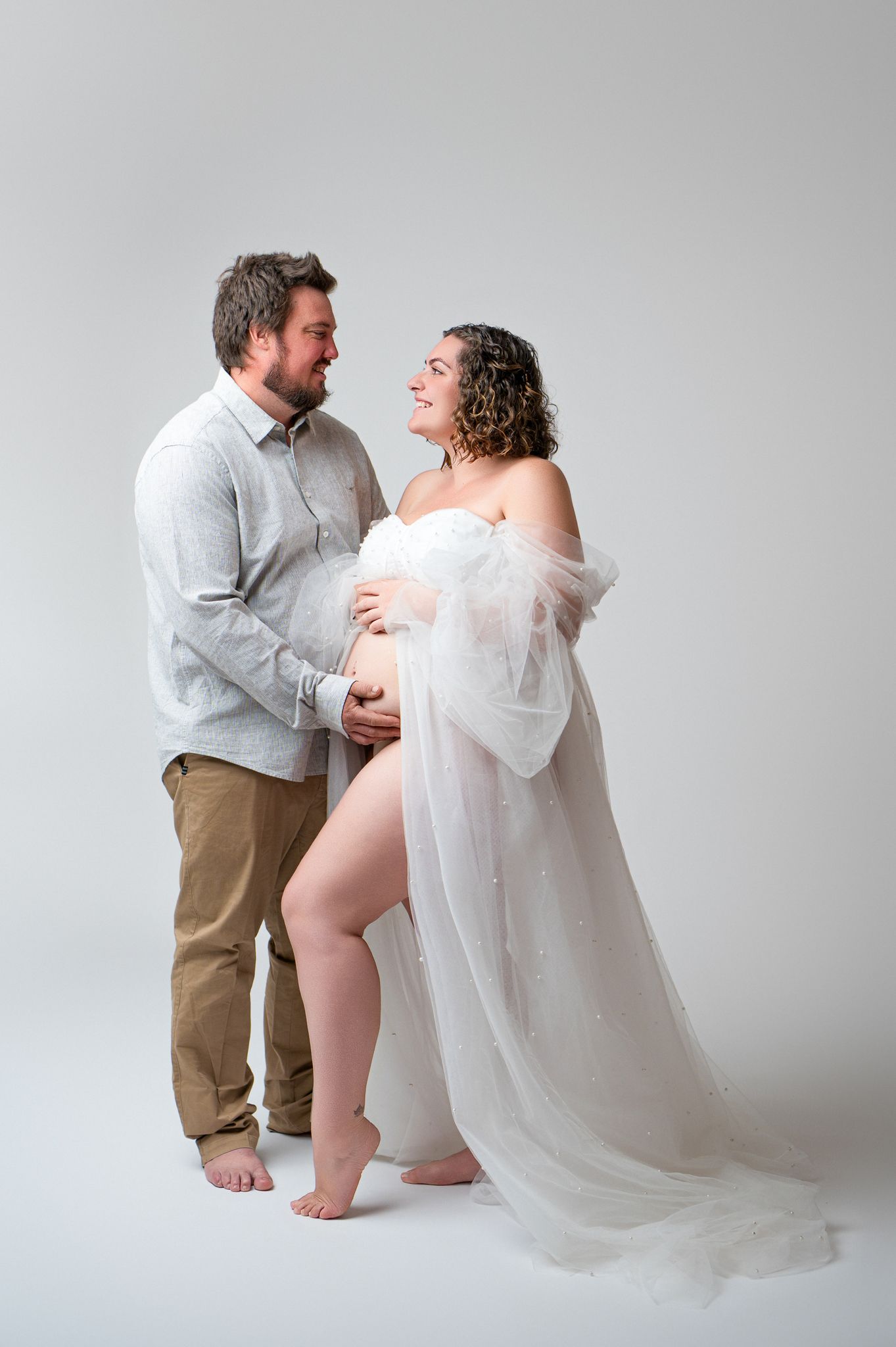 Couple sharing a tender moment during a maternity photo shoot, woman in white sheer gown cradling baby bump.