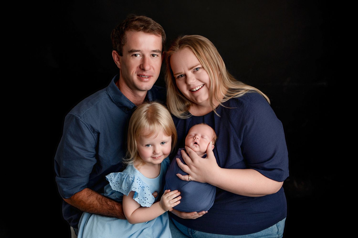 Family of four posing for professional portrait, parents with toddler girl and newborn baby wrapped in blue, black background.