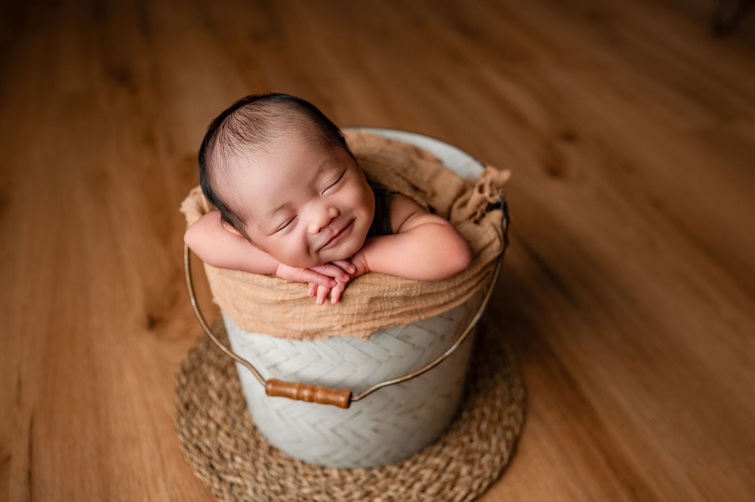 Smiling newborn baby sleeping peacefully in a rustic bucket with burlap lining on a wooden floor.