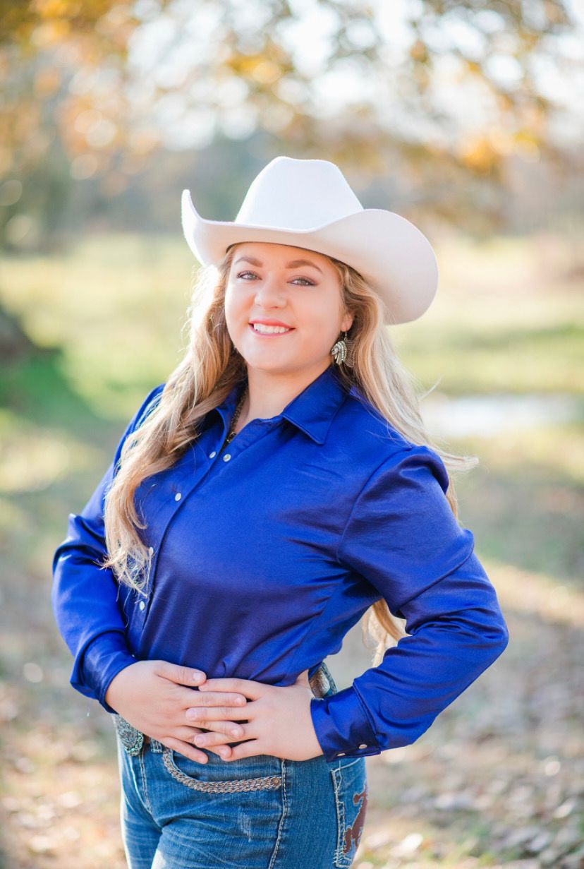 A person in a bright blue button-up shirt and white cowboy hat poses outdoors with a natural autumn background.