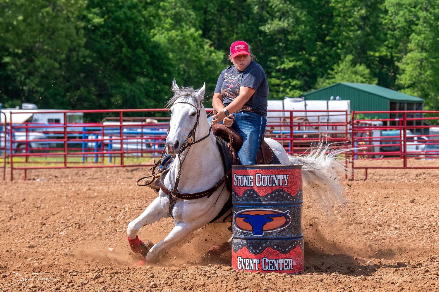 A rider steers a gray horse around a barrel in a dirt rodeo arena with red fencing during a competitive barrel racing event.