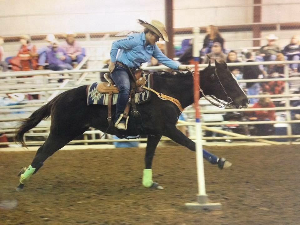 A rodeo competitor rides a black horse through pole bending competition in an indoor arena.