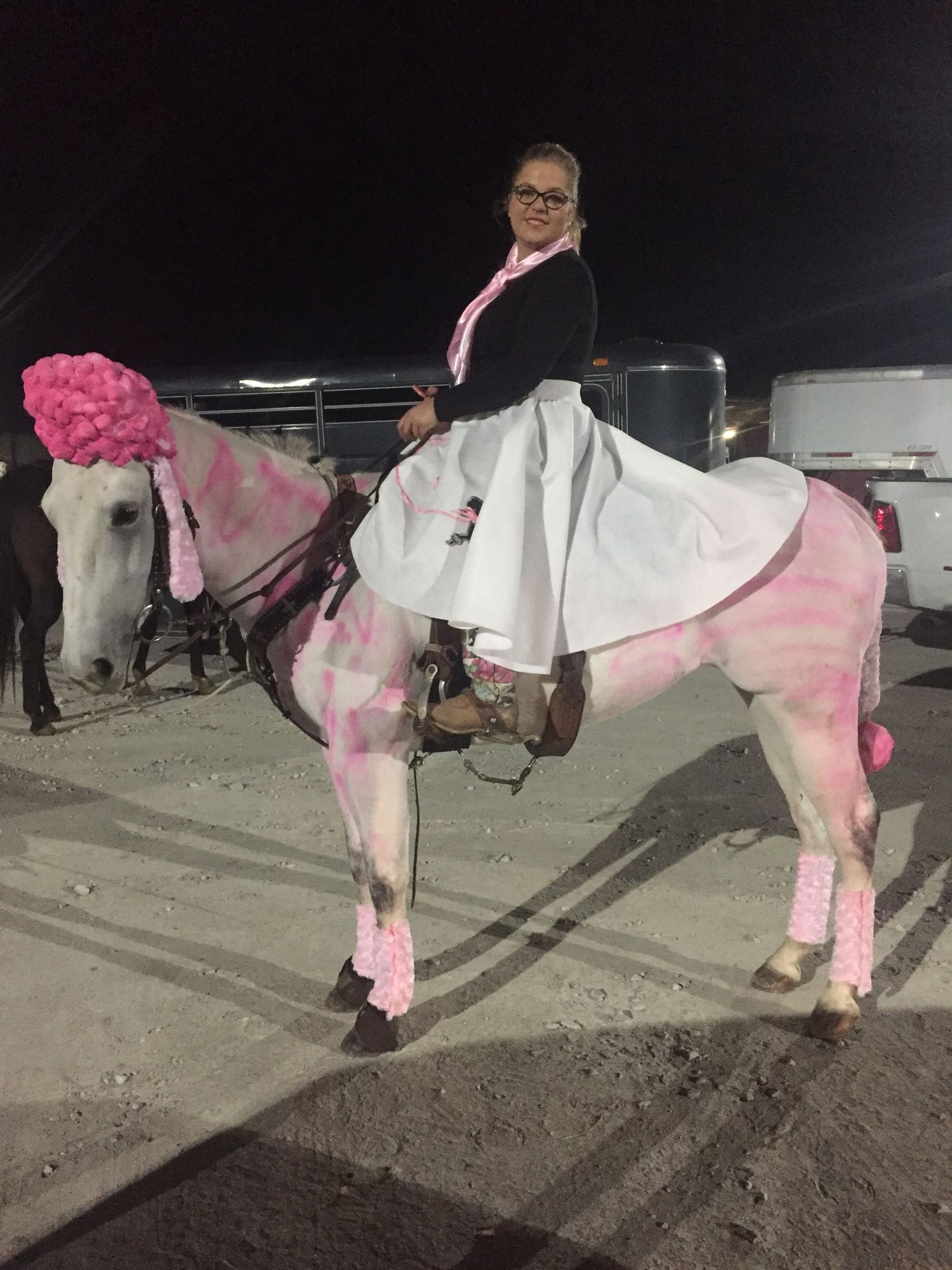 A white horse decorated in pink accessories and paint stands with a rider in a dark parking lot at night.