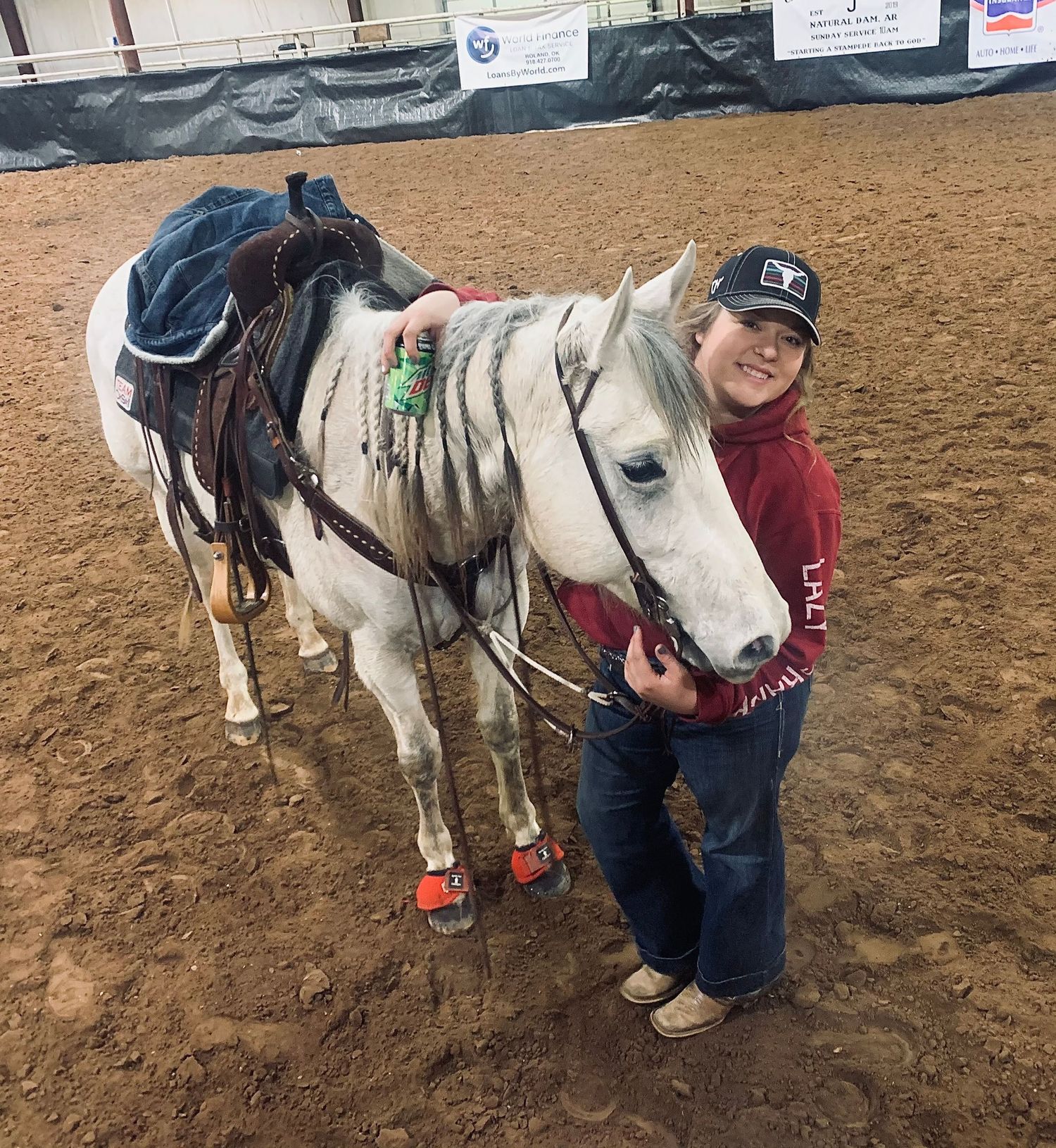 A white horse with red boots and saddle stands in a dirt arena alongside someone in a red jacket.