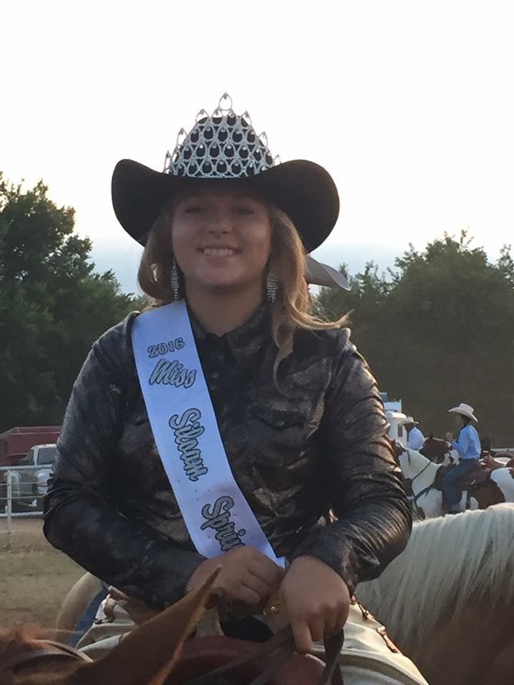 A rodeo rider on horseback wearing a black cowboy hat, crown, and show attire with a sash in an outdoor arena at dusk.