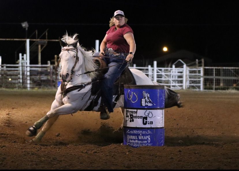 Barrel racing competitor on white horse gallops around blue barrel in nighttime rodeo arena during competition.