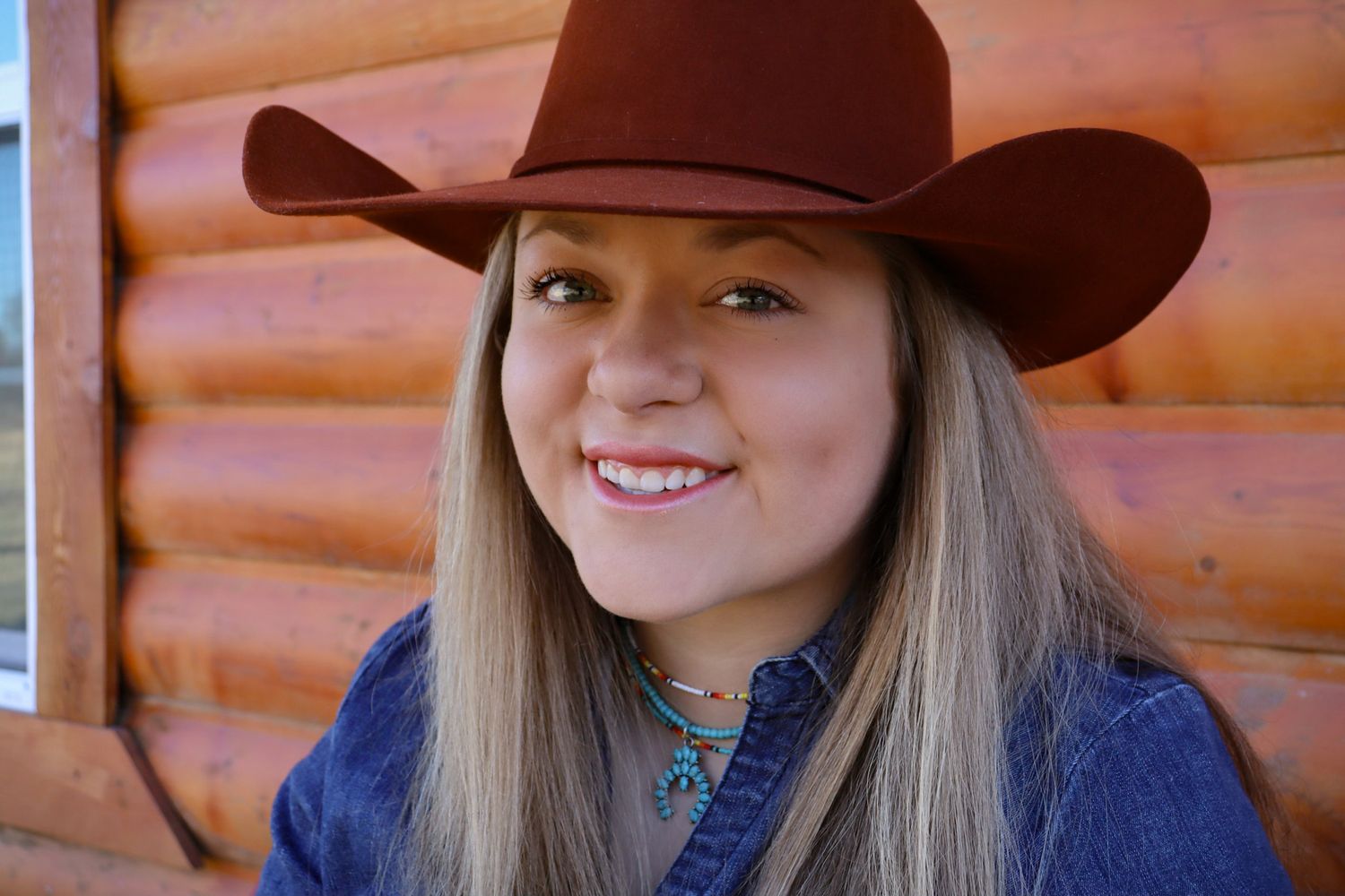 A western-style portrait showing someone in a brown cowboy hat and denim shirt against a rustic log cabin background.