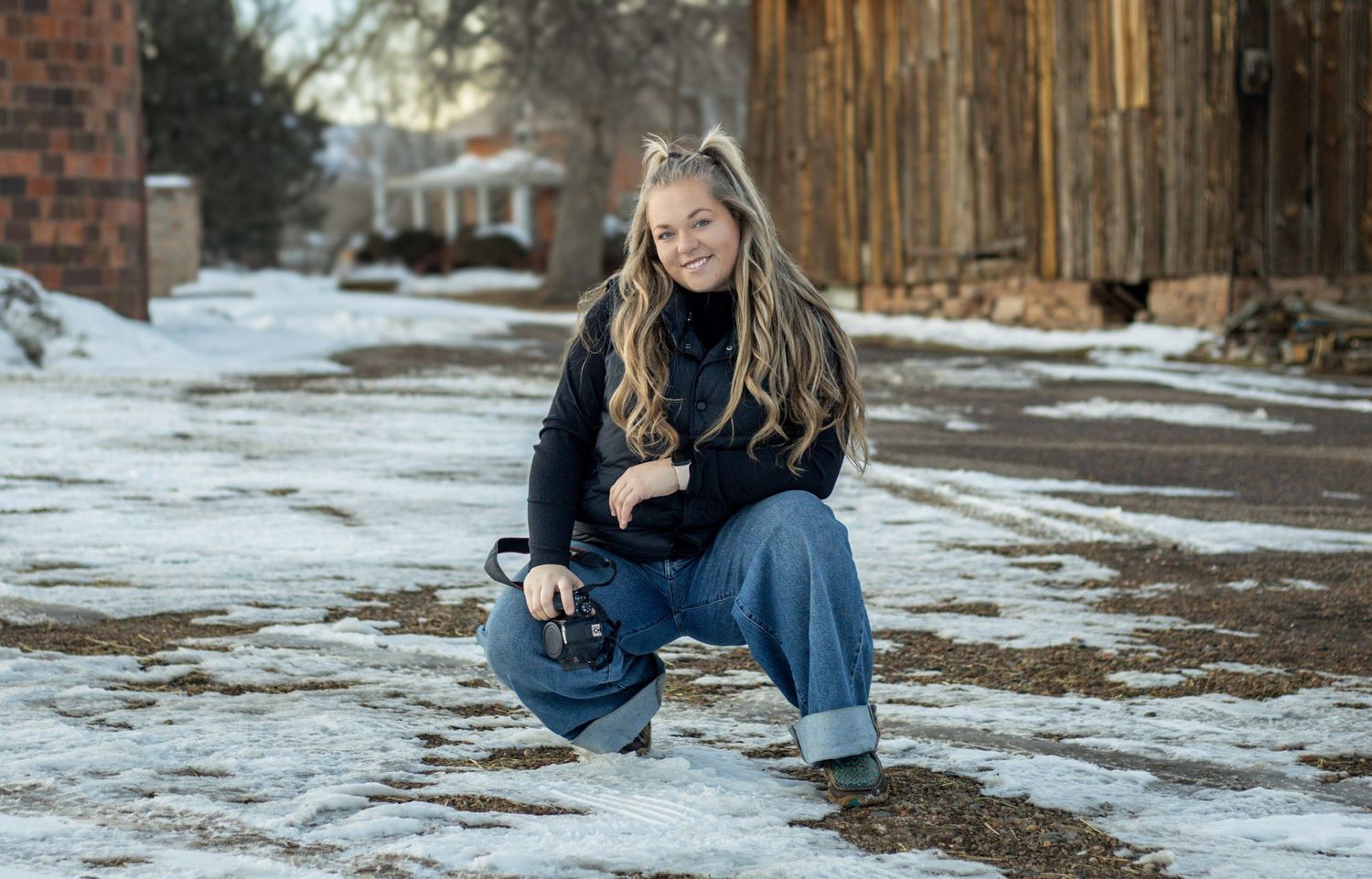 Young blonde woman crouching in snowy alley holding camera, wearing jeans and black sweater, winter outdoor photography session.