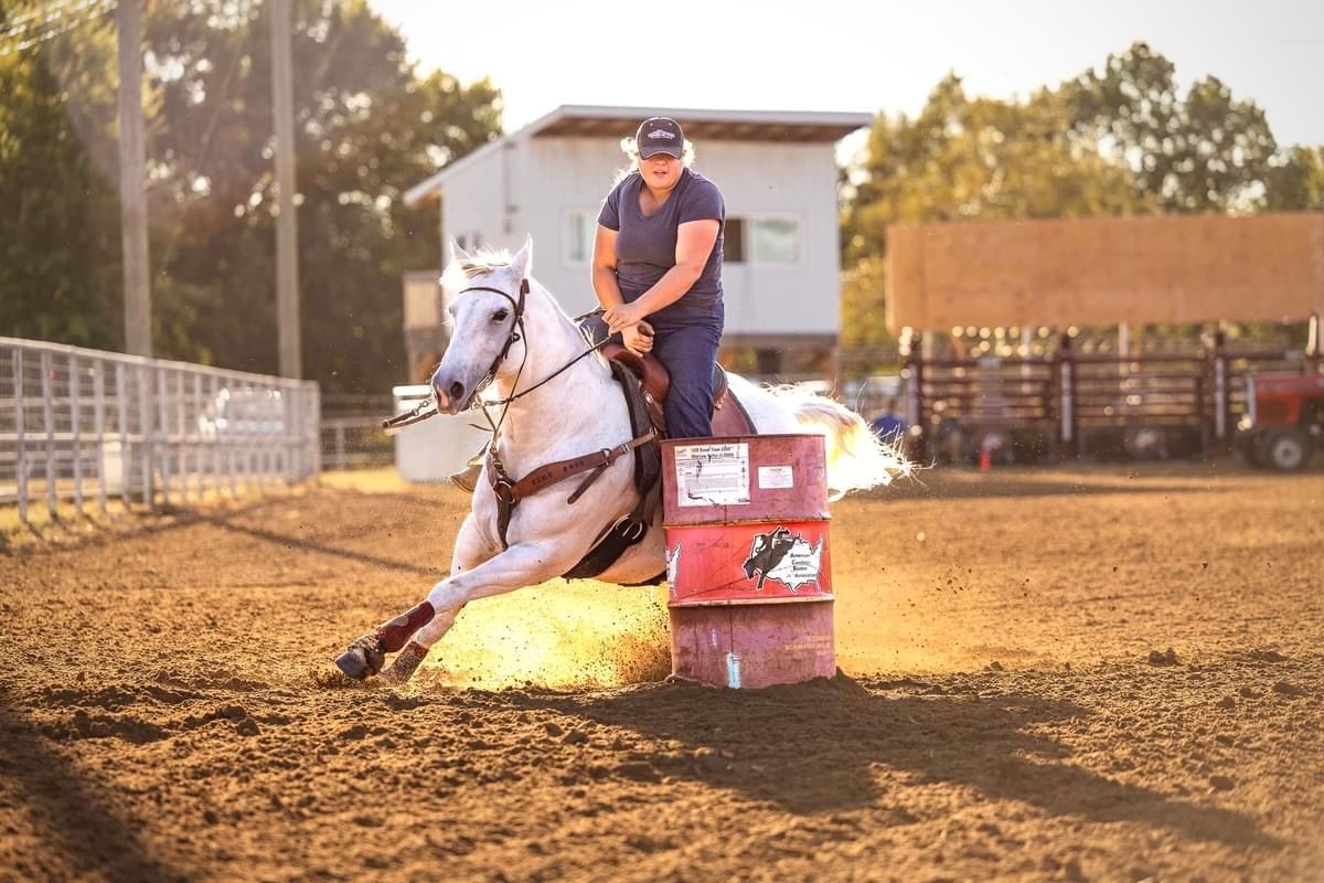 A rider and horse navigate around a barrel during a competitive rodeo event at sunset in a dusty arena.