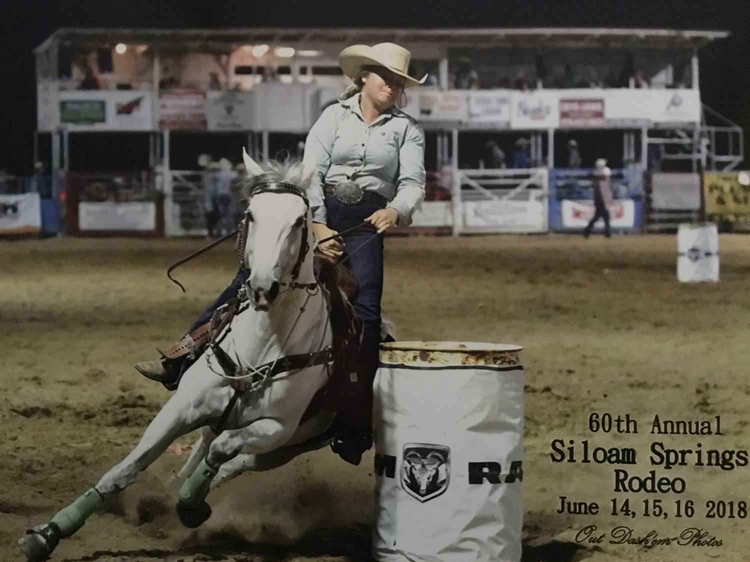 A cowgirl barrel racing on horseback at nighttime rodeo competition event with stands in background.