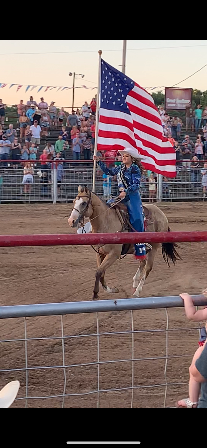 A horse rider carrying an American flag performs during a rodeo event as spectators watch from crowded bleachers.