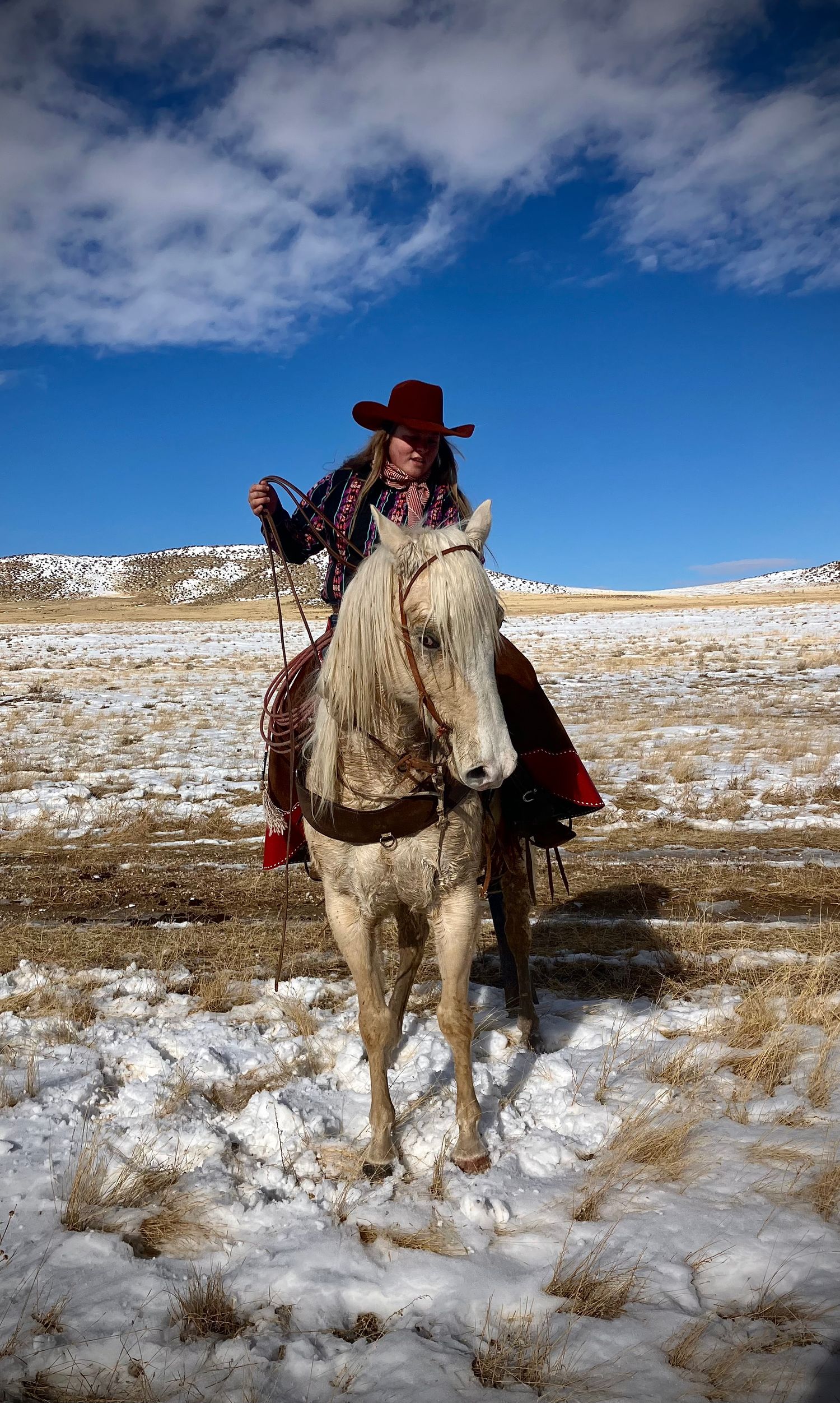A horse rider in western attire traverses a snowy winter landscape under bright blue skies with white clouds.