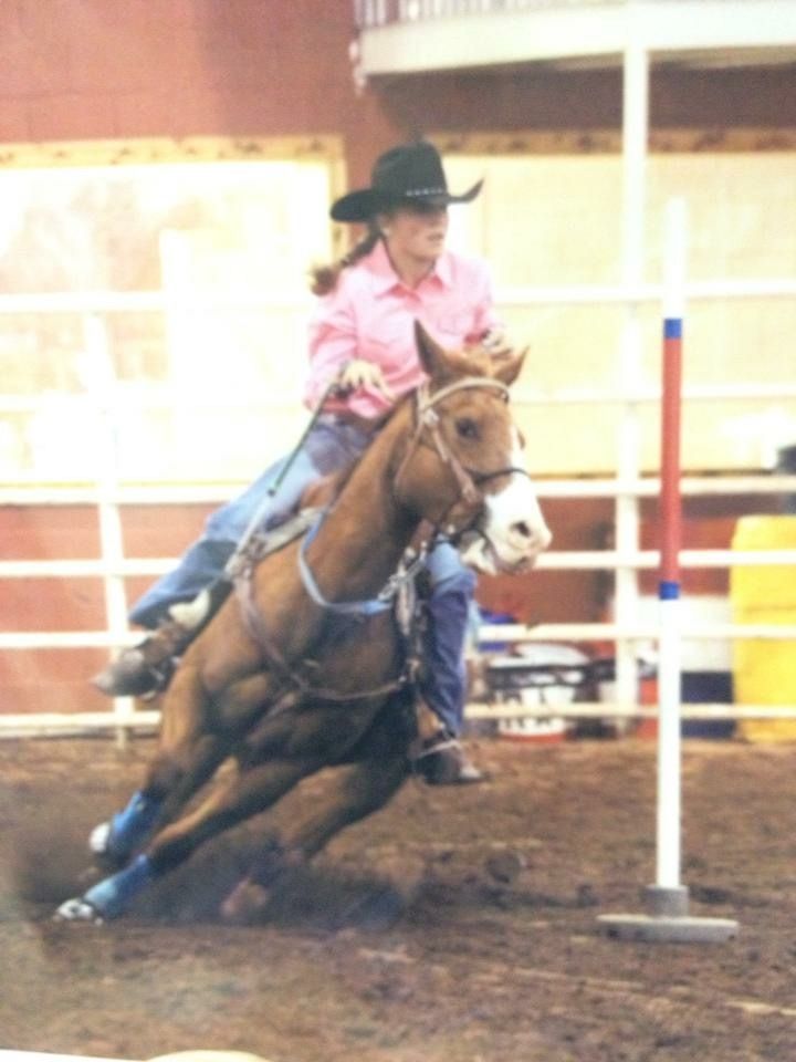 A rider in a pink shirt and cowboy hat competing in barrel racing on a brown horse around a pole in an arena.