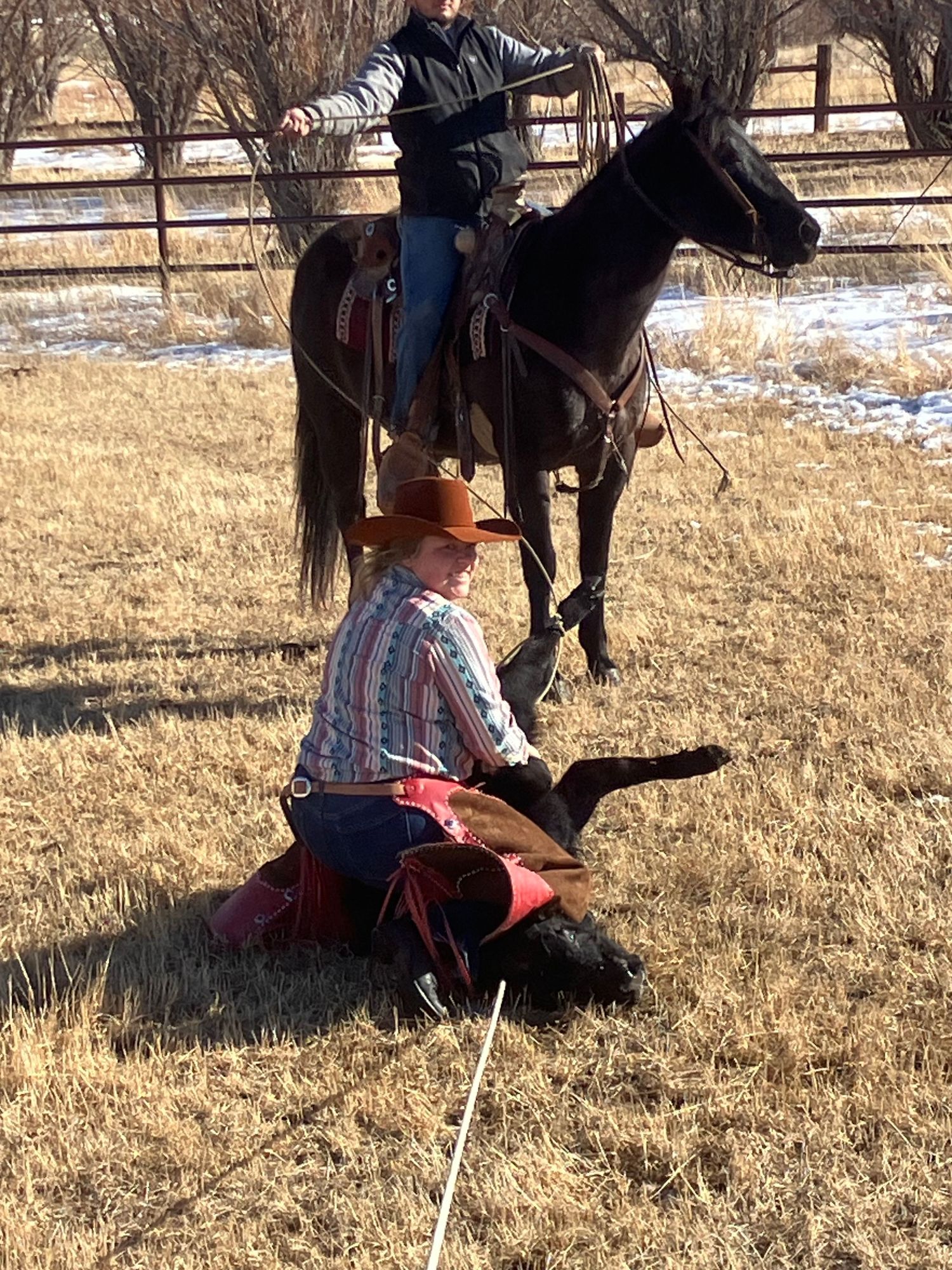 Winter scene with horseback rider and cowgirl sitting in snowy field next to brown fenced corral.