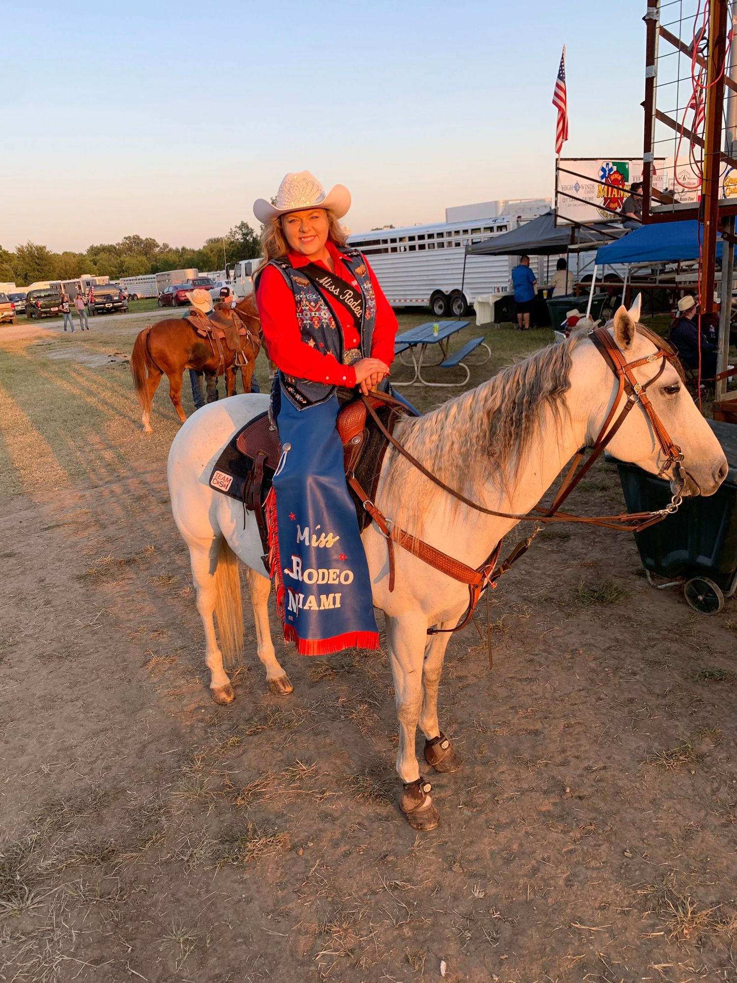 A rider in red western attire sits atop a light-colored horse at a dusty outdoor event with an American flag in view.