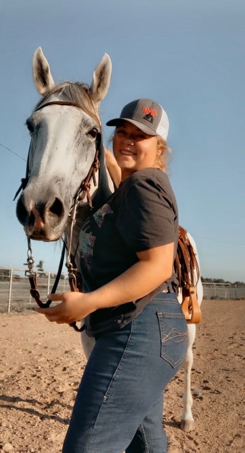 A person in jeans and baseball cap stands beside a white horse with bridle in a sandy outdoor paddock.