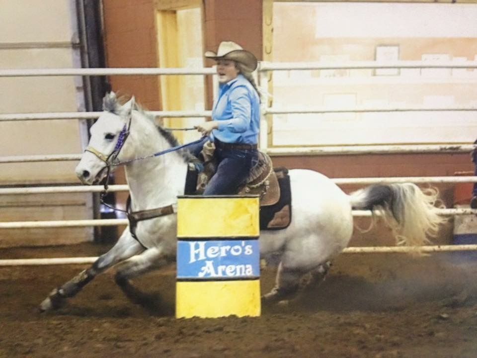 A rider on a white horse navigates around a bright yellow barrel in an indoor arena during a barrel racing competition.