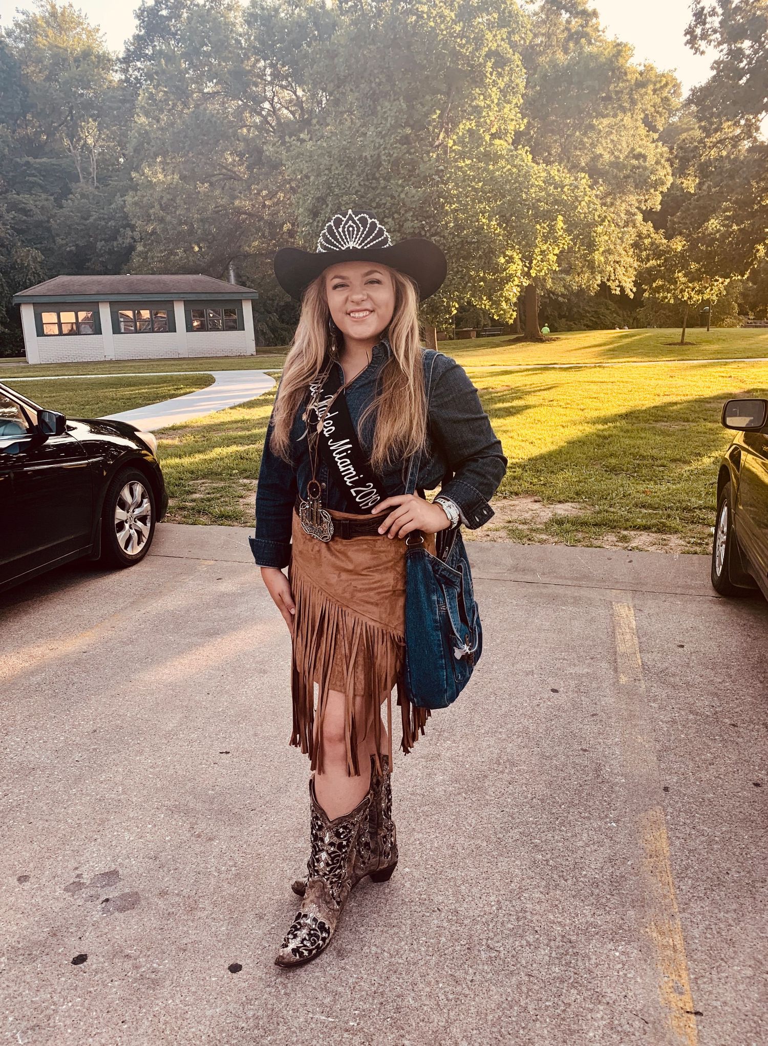 A person wearing a cowboy hat, fringe suede skirt and snakeskin boots poses outdoors at sunset in a parking lot.