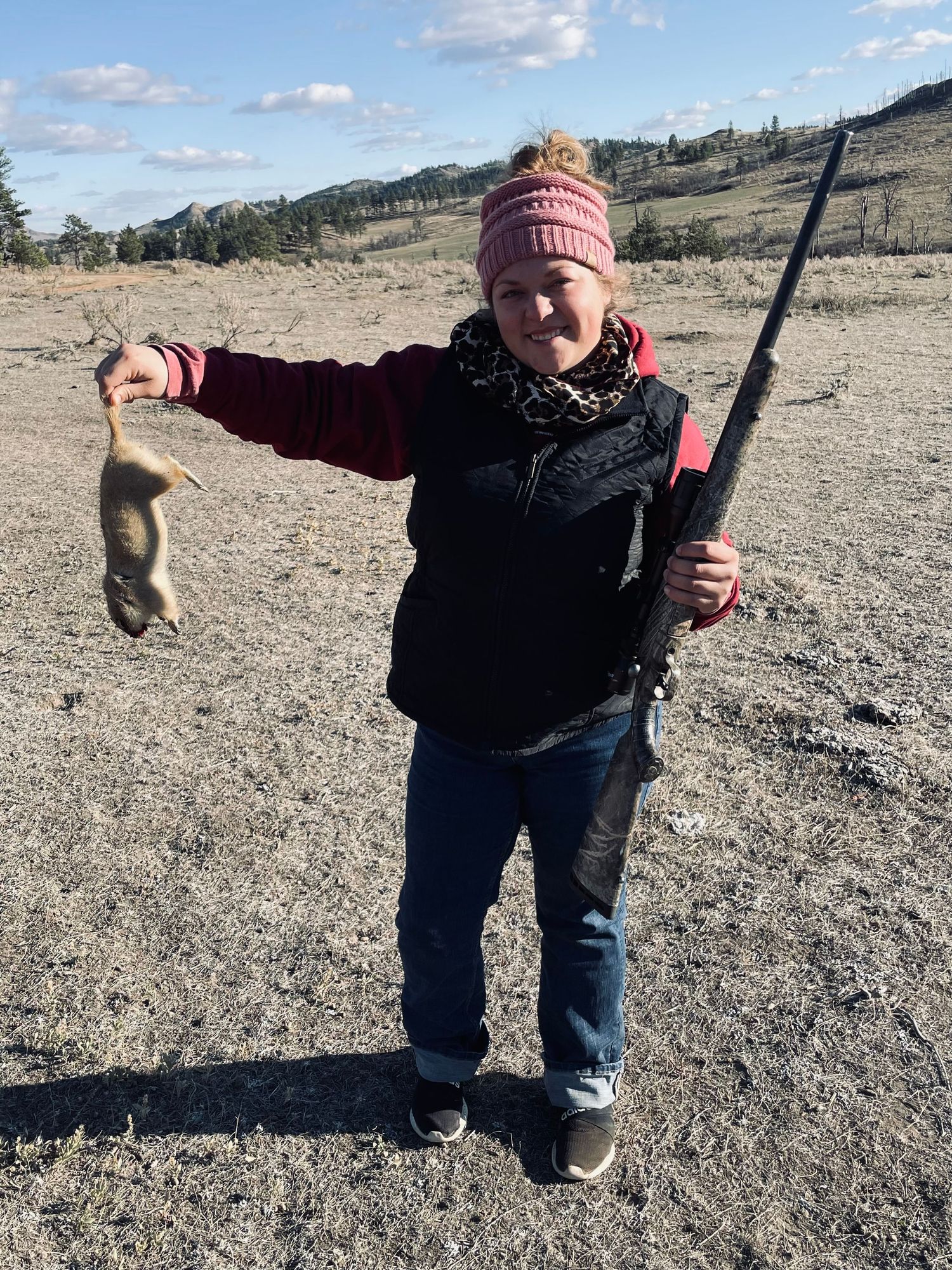 A young hunter in a pink beanie and black coat holds up their catch and fishing rod in a mountainous winter landscape.