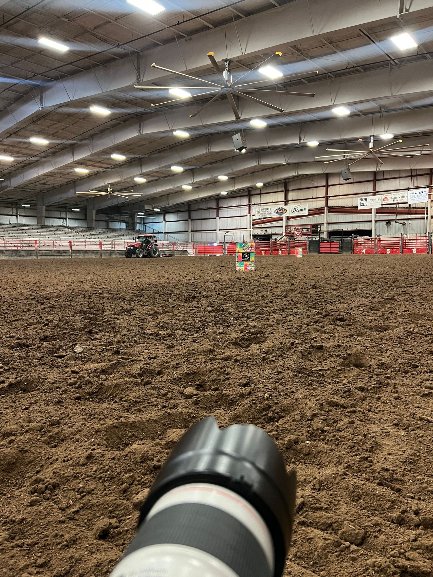 View of an indoor equestrian arena with dirt floor and fluorescent lighting seen through a camera lens in the foreground.