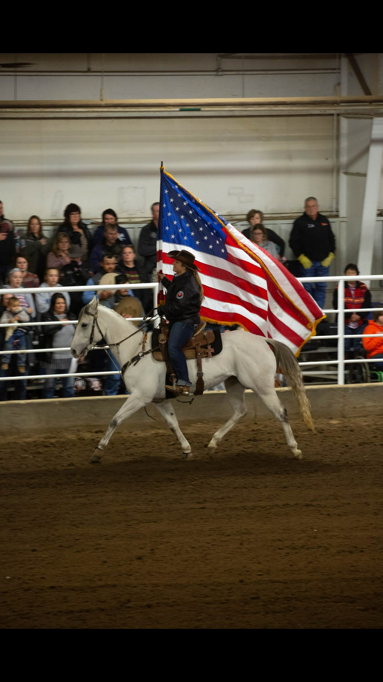 A rider carries the American flag while riding a white horse in an indoor arena with spectators in the background.