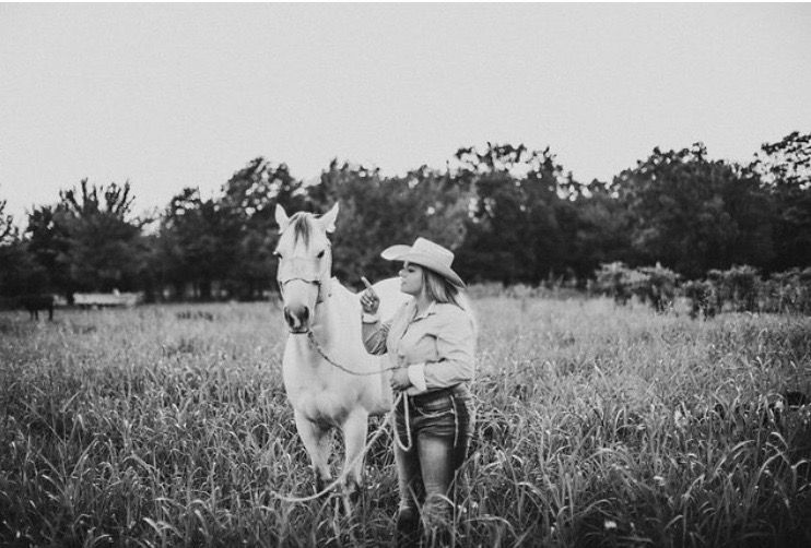 A black and white photograph of someone in cowboy attire standing with a white horse in a grassy field.