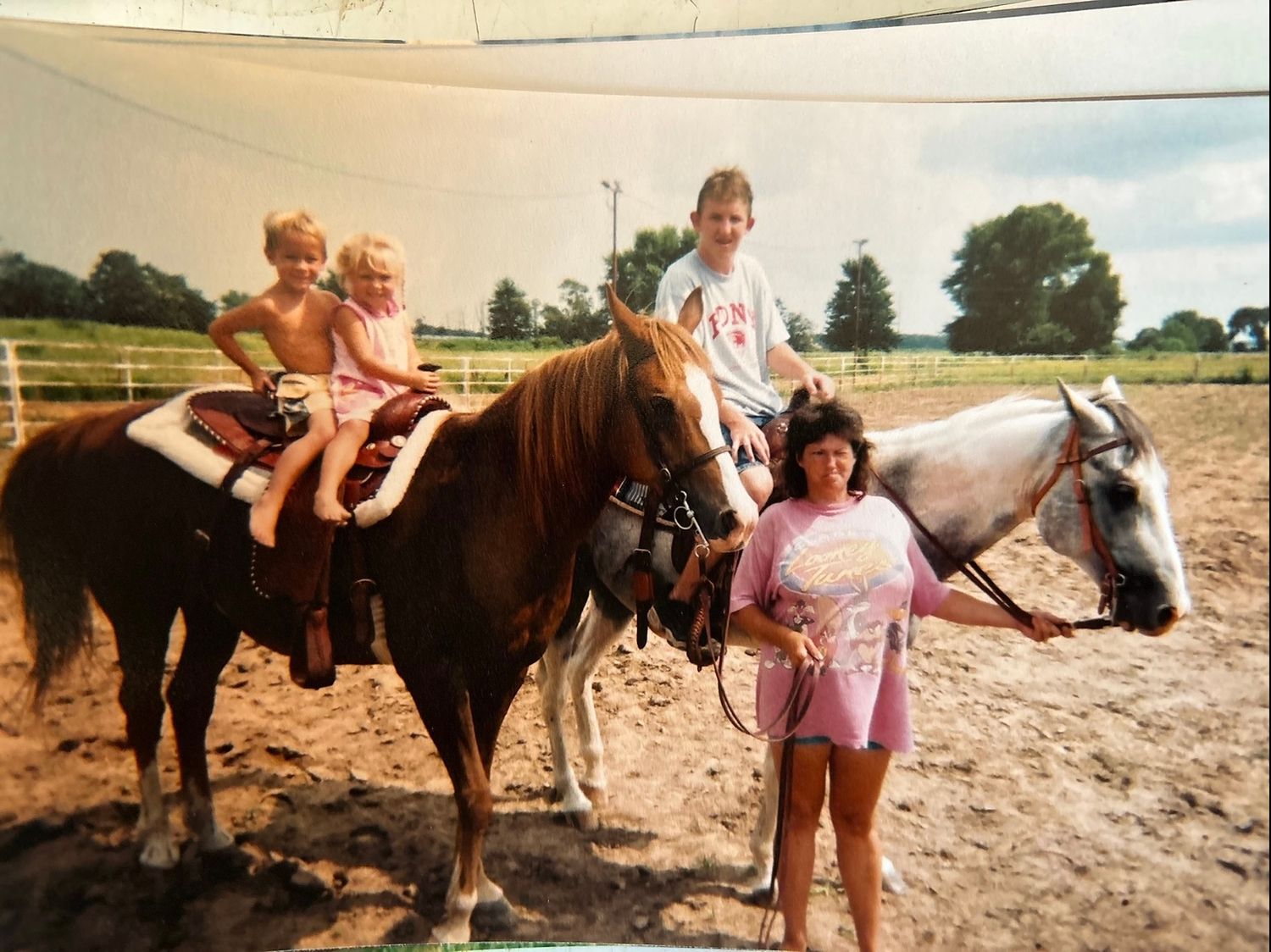 Horseback riding on two horses in a sandy corral with ranch fencing and trees in the background.