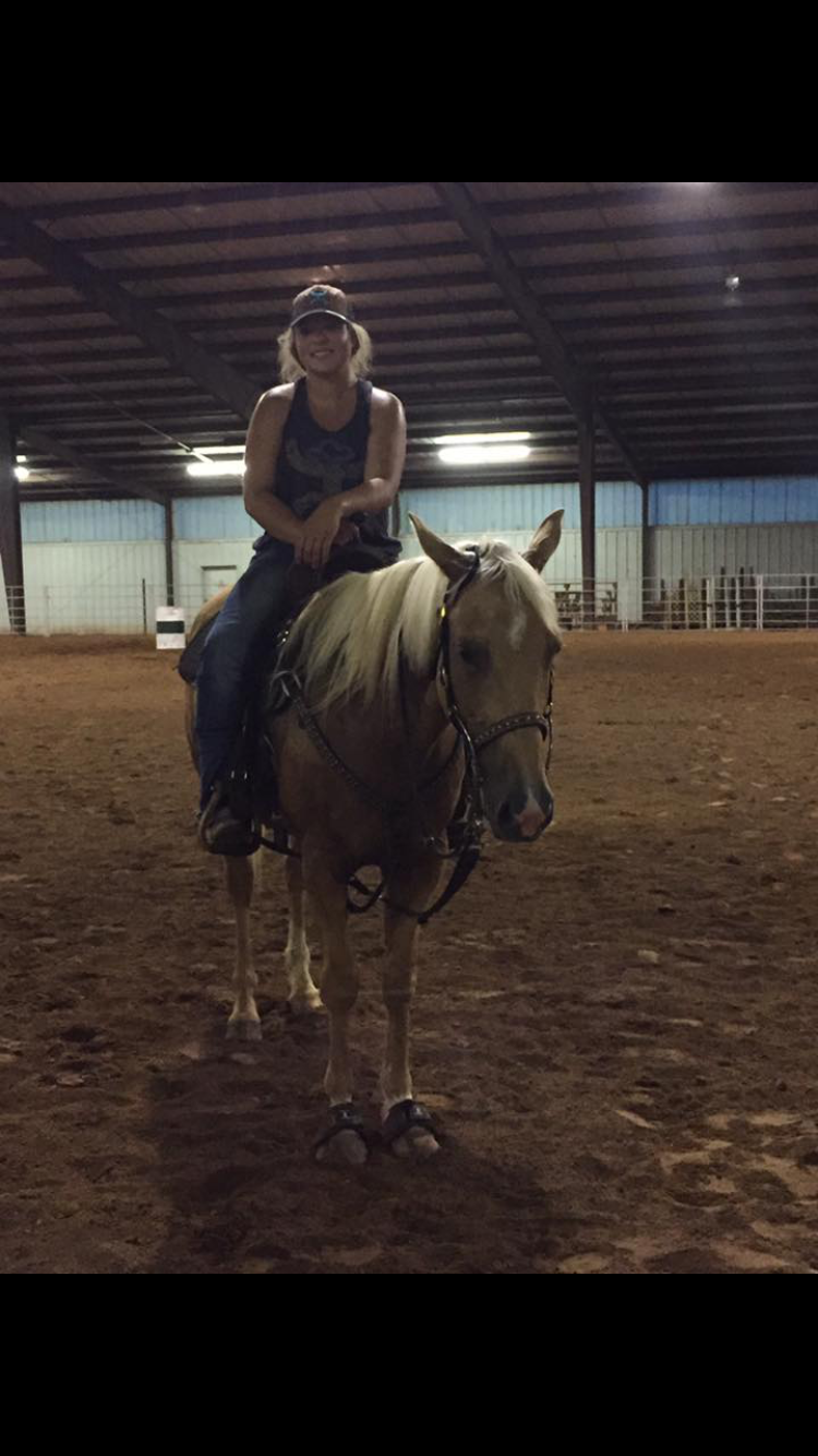 A rider sits atop a light-colored horse inside an indoor riding arena with dirt flooring and exposed beams overhead.