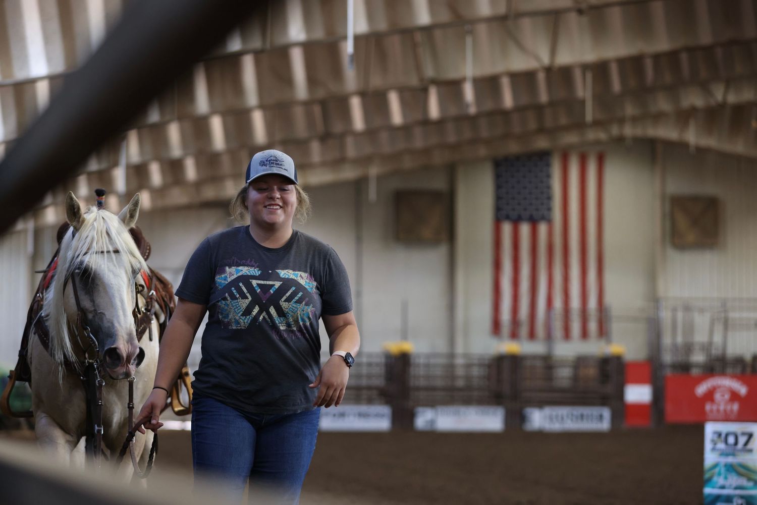 A person wearing a gray t-shirt walks past a large American flag hanging inside a barn or arena.