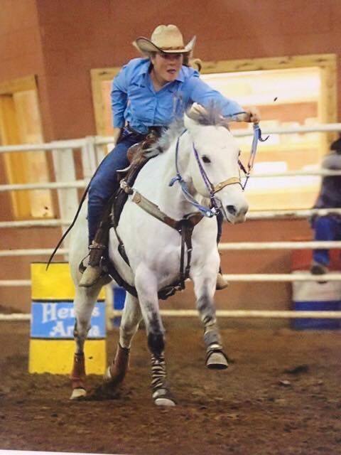 A rider in a blue shirt and cowboy hat guides a white horse around a barrel during a rodeo competition.