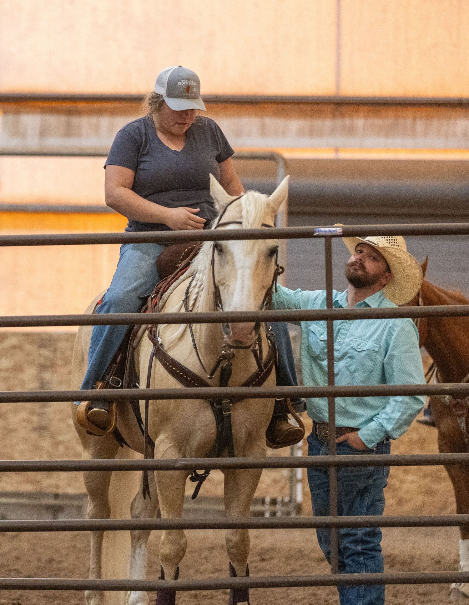 Woman on white horse in arena receives instruction from cowboy in turquoise shirt and hat.