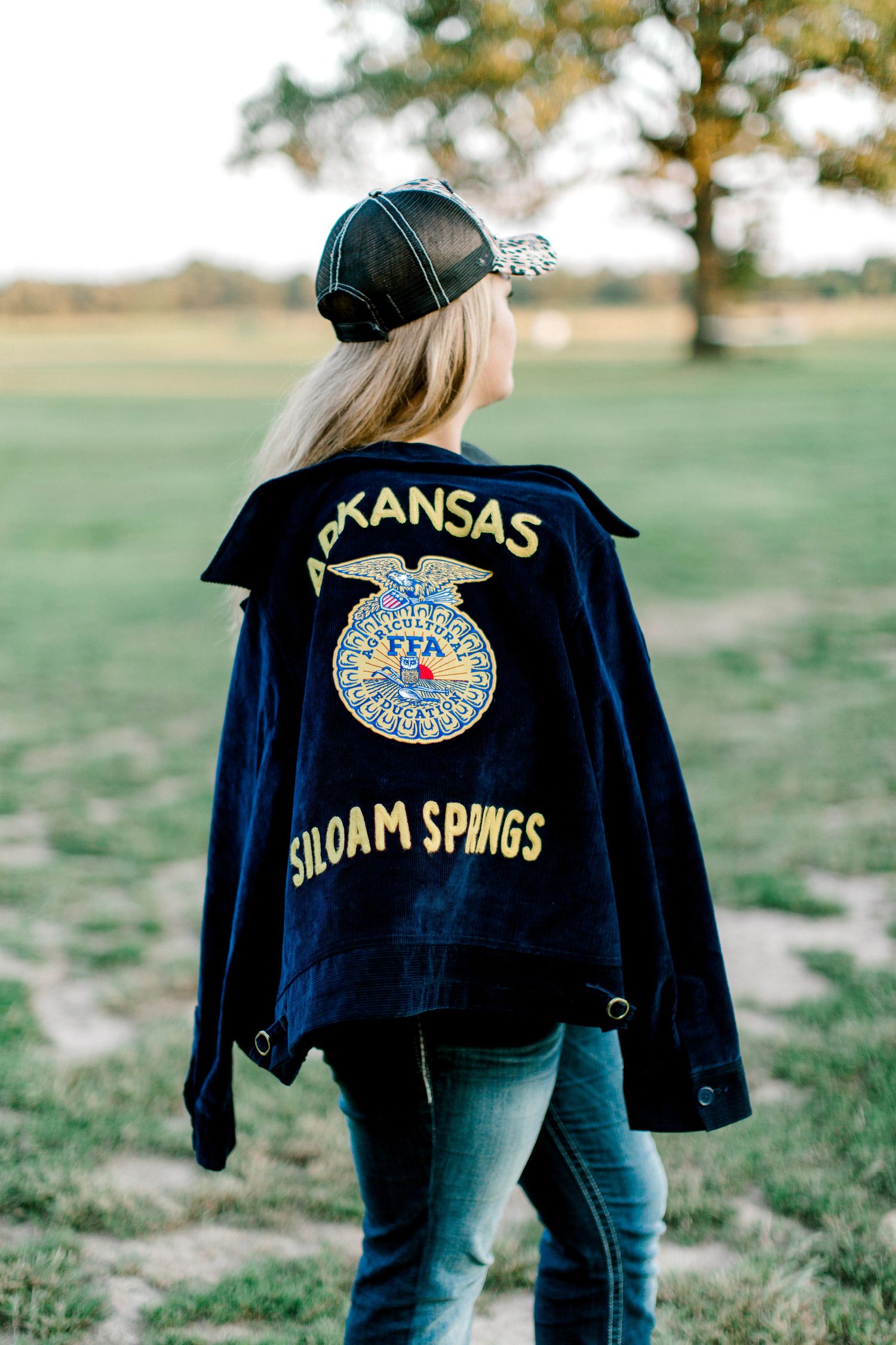 A person wearing a Kansas FFA Siloam Springs jacket and baseball cap stands in an open field.