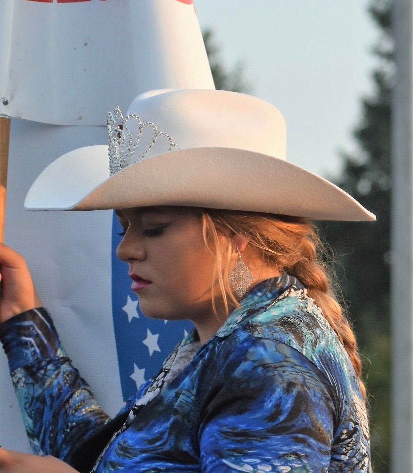 A person in a blue jacket wears a silver tiara and light-colored cowboy hat against a blurred outdoor backdrop.
