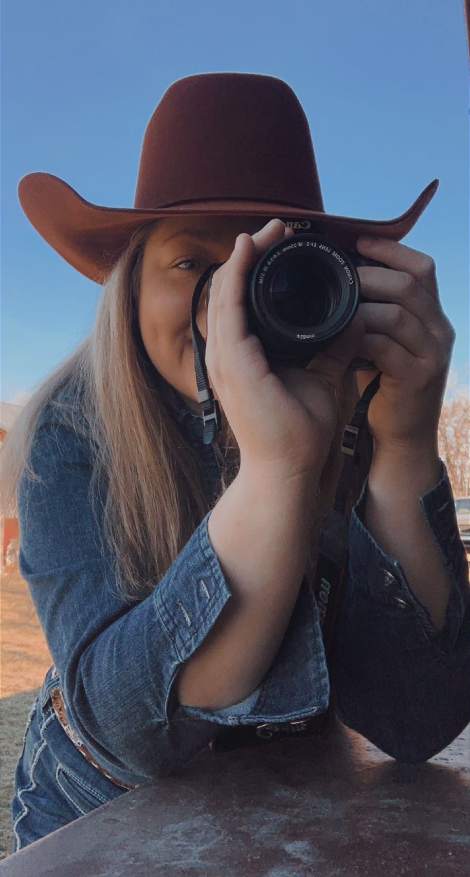 A photographer wearing a brown cowboy hat and denim shirt takes a photo with a digital camera against a blue sky.