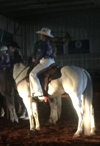 A cowboy rides a white horse illuminated by spotlights in a dimly lit indoor arena.