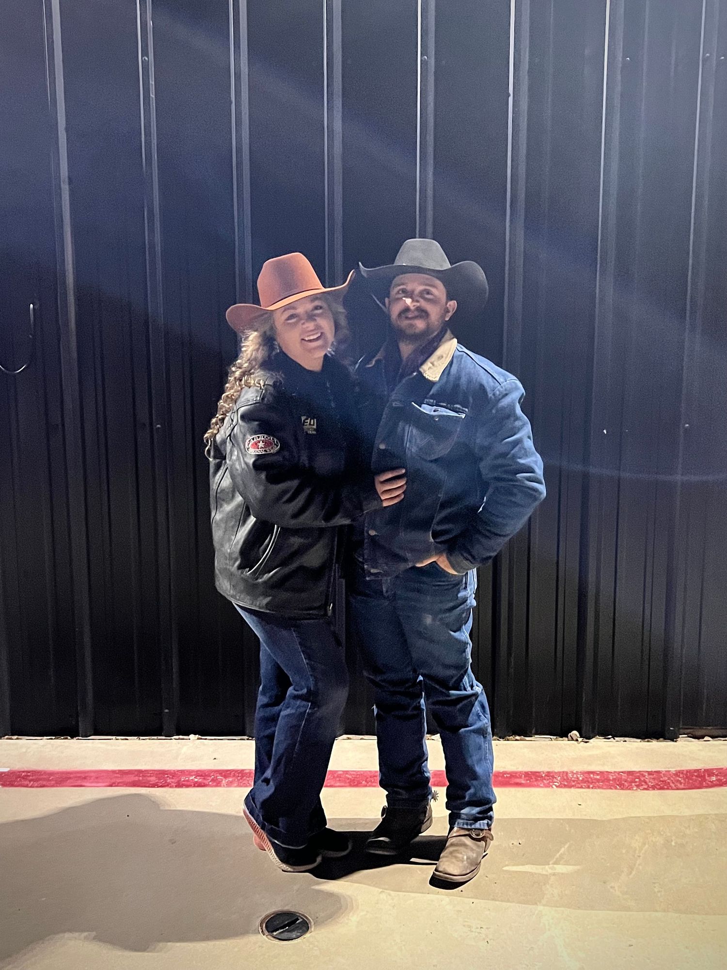Two individuals wearing cowboy hats and western attire pose together against a dark metal wall.