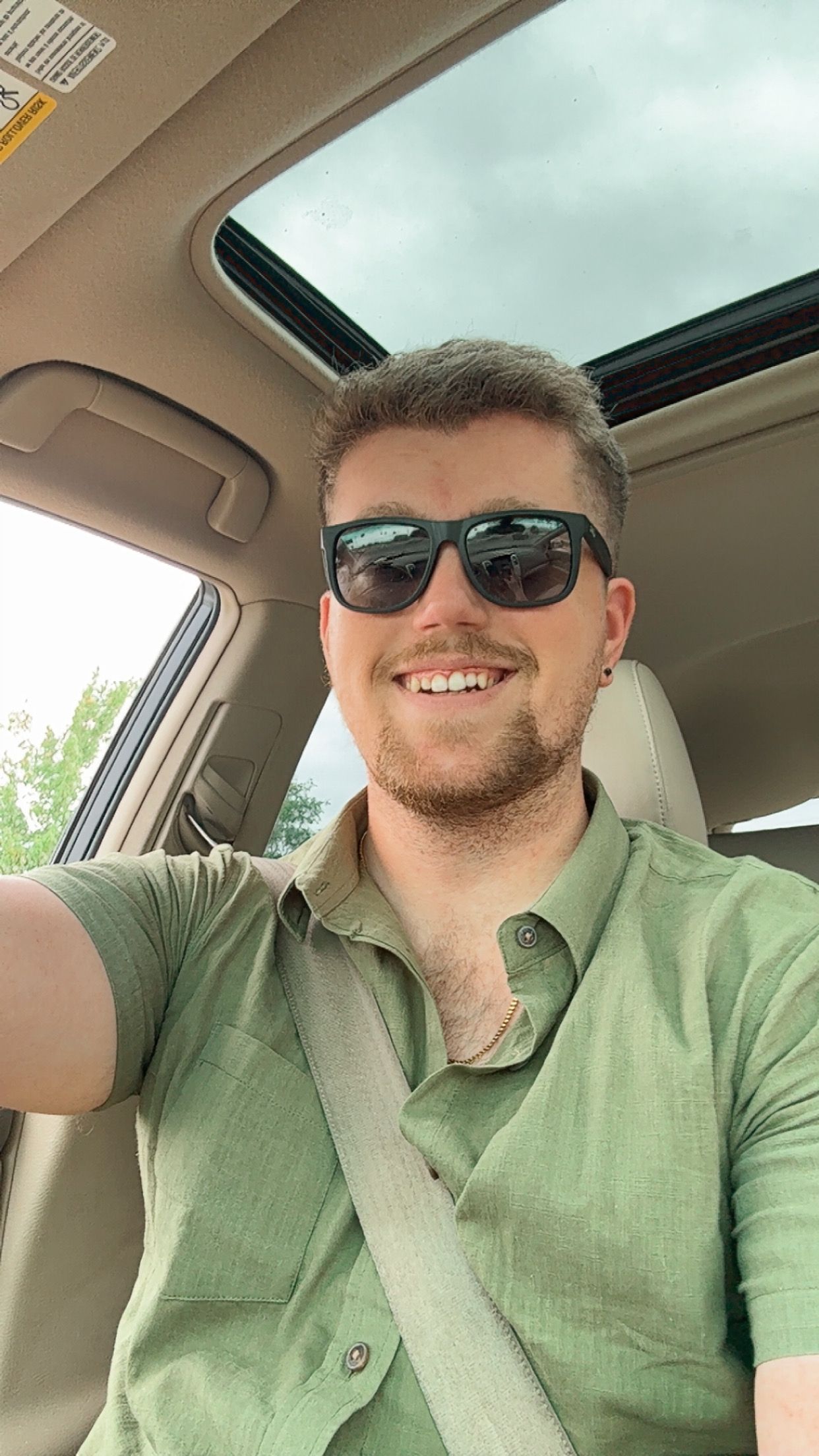 Smiling man wearing sunglasses and green shirt taking a selfie in a car with sunroof, seatbelt on.