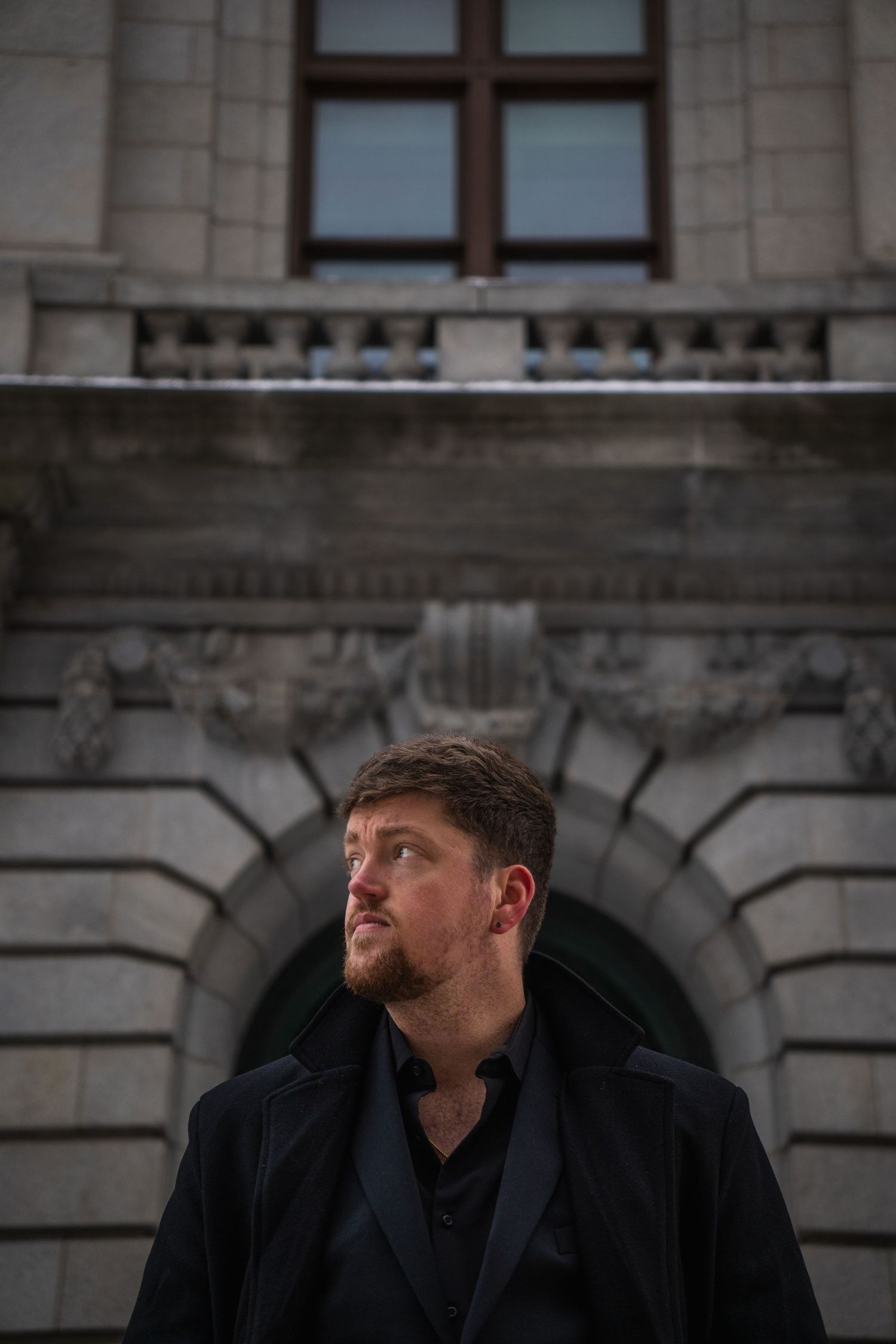A bearded man in a black coat gazes upward before a grand stone building with arched doorway.