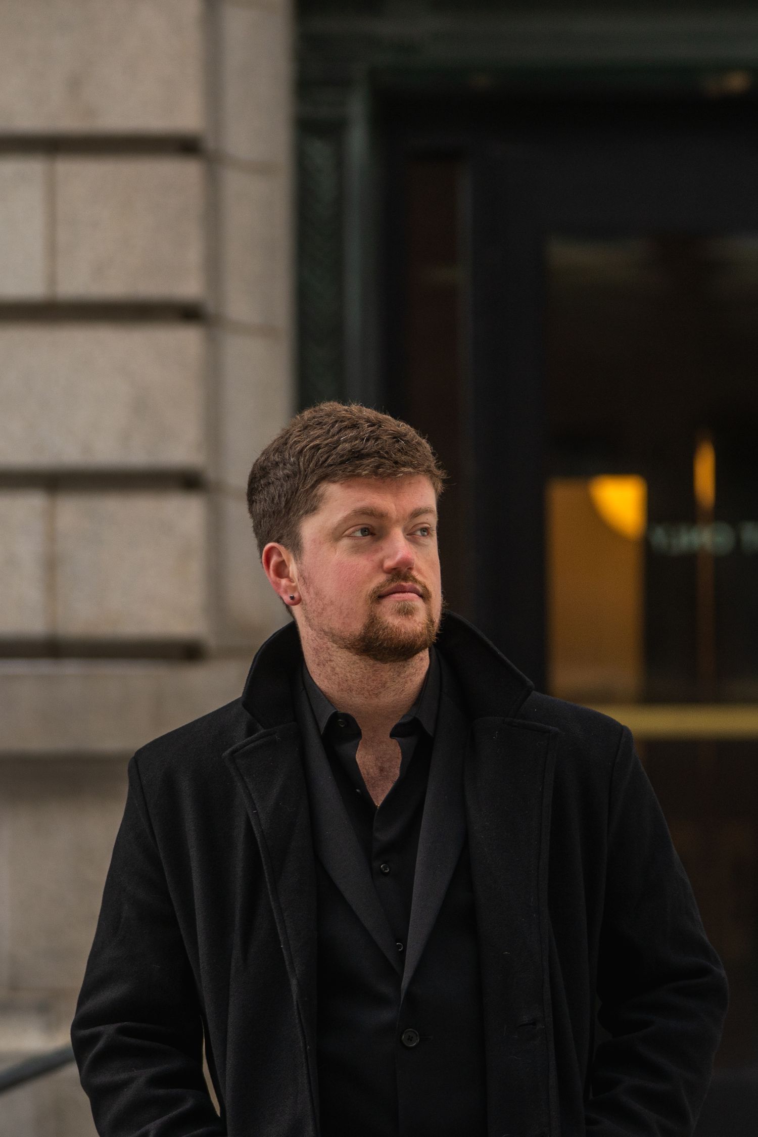 Man in black coat and shirt standing outdoors near a building, looking thoughtfully to the side.