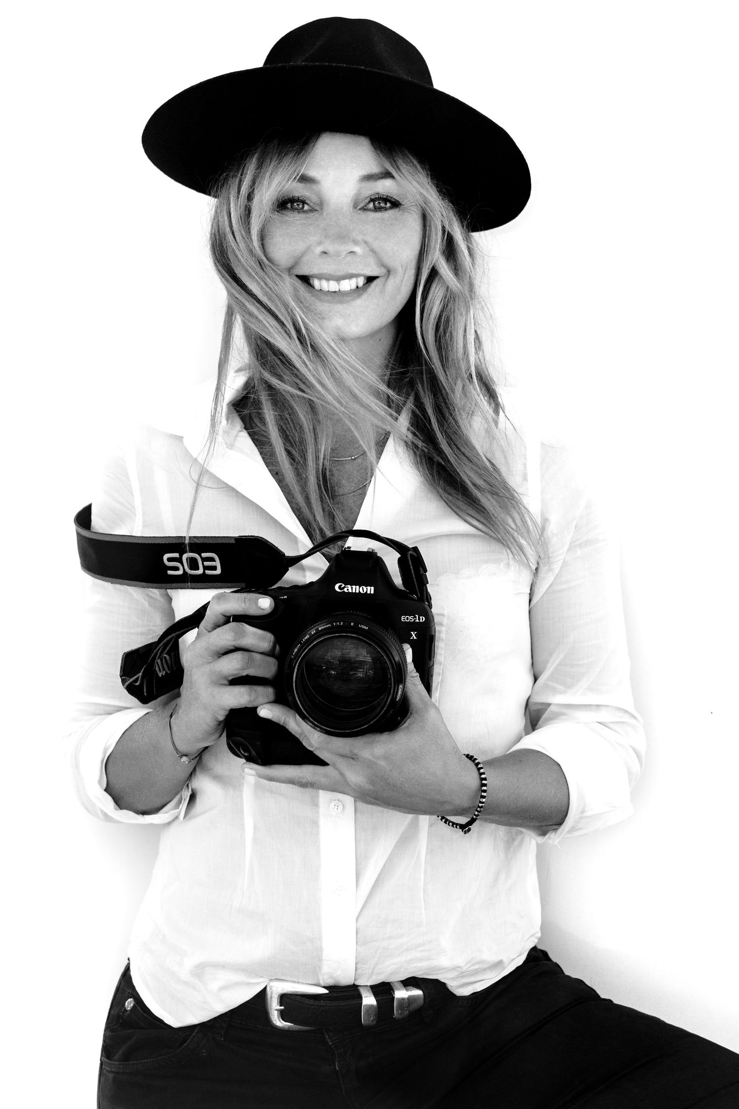 Black and white portrait of a photographer wearing a black hat and white shirt holding a Canon camera and smiling.