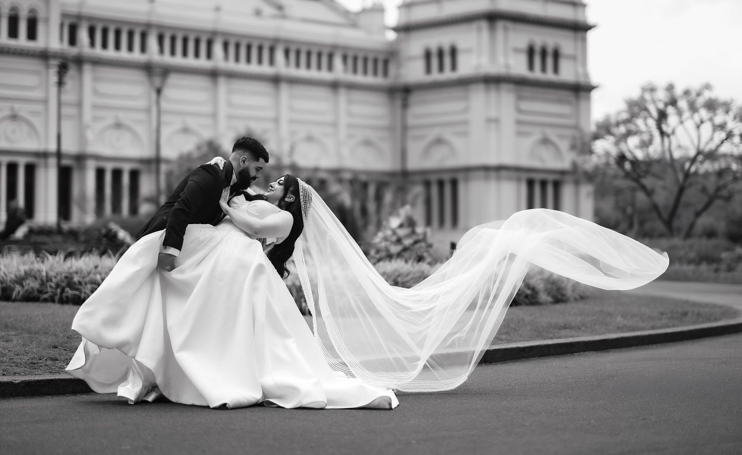 A long flowing white wedding dress and veil trail dramatically across a paved walkway in black and white photography.