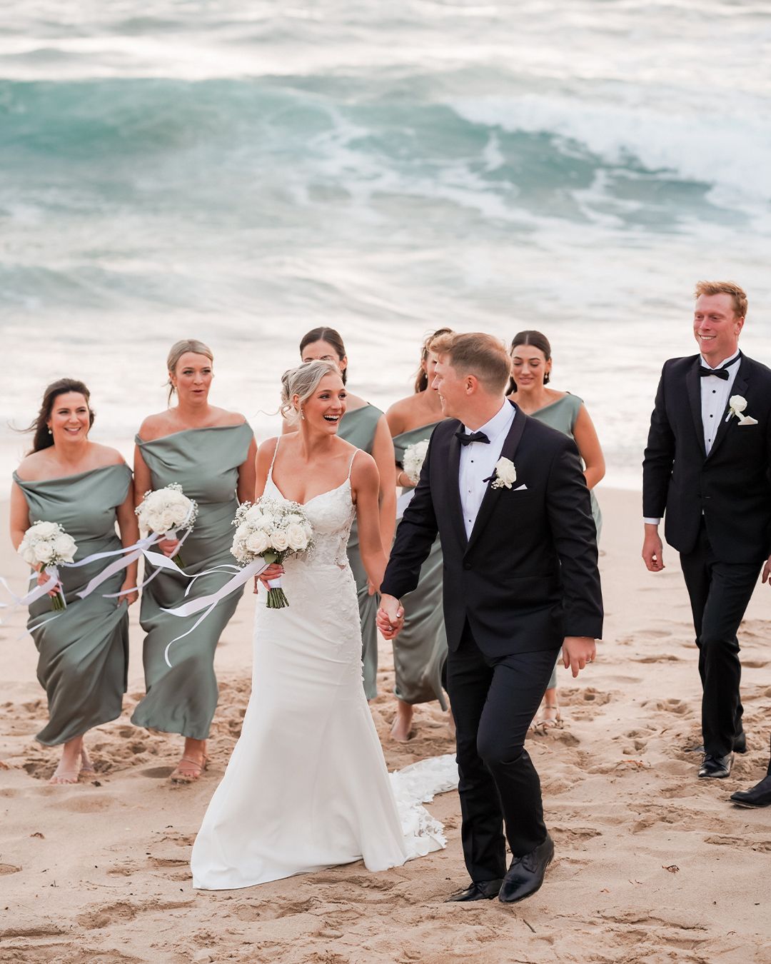 Bride and groom walk hand in hand on beach with bridesmaids in sage green dresses and groomsmen in black tuxedos.