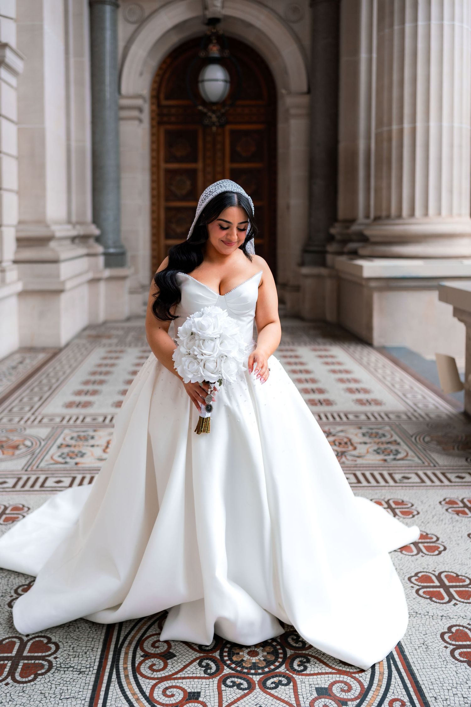 Bride in elegant white ball gown poses gracefully on ornate tiled floor in grand architectural hallway.